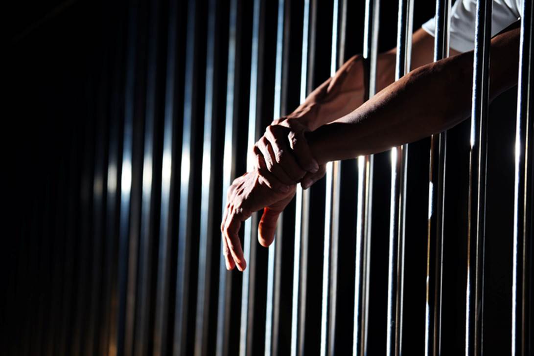 A prisoner’s hands grasp the bars of a cell. This image is used in an article discussing false imprisonment and federal law, alluding to a situation of wrongful incarceration.